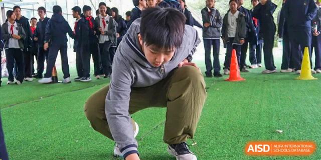 A male student crouches down on an artificial turf field, reaching out his hand, perhaps marking a spot or picking up an item, at an outdoor sports practice at Hangzhou Entel Foreign Language School.