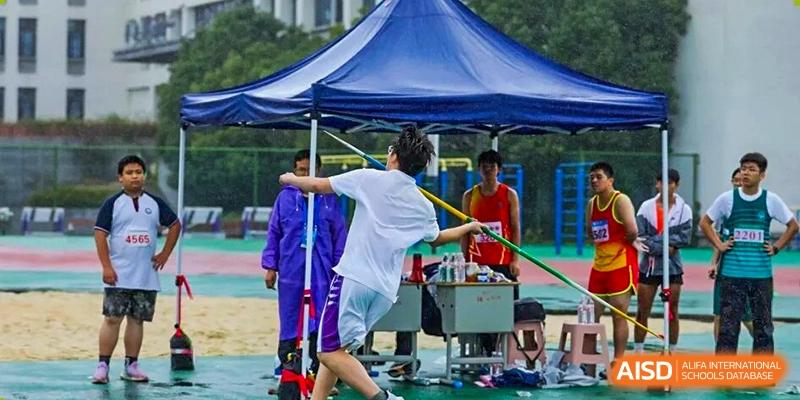 A student throws a discus or similar object in an outdoor event near a tent, with staff and others nearby at Hangzhou Entel Foreign Language School.