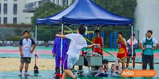 A student throws a discus or similar object in an outdoor event near a tent, with staff and others nearby at Hangzhou Entel Foreign Language School.