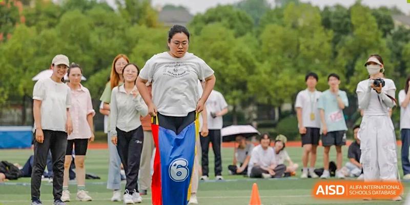 A male student competes in a sack race, wearing a large sack up to his waist, while other participants and spectators watch at Hangzhou Entel Foreign Language School.