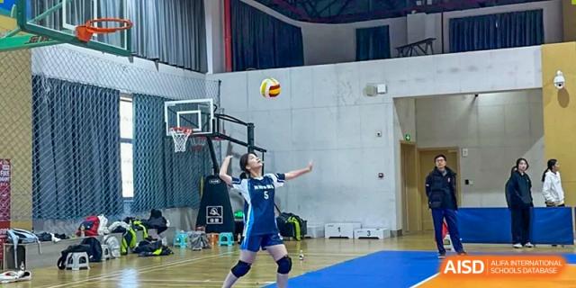 A determined student from Hangzhou Entel Foreign Language School performing an overhead serve during an indoor volleyball game in the gymnasium.
