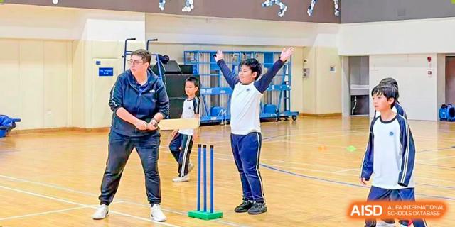 A teacher supervises students practicing cricket drills with wickets set up indoors at BASIS International School Park Lane Harbour.