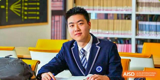 A young male student in a dark blazer and tie smiles confidently at the camera while sitting in a library at BASIS International School Park Lane Harbour.