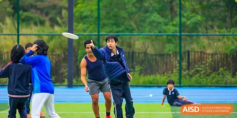 Two young men are reaching up to catch a flying disc during an intense game of Ultimate Frisbee outdoors at BASIS International School Park Lane Harbour.
