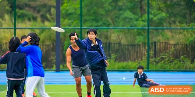 Two young men are reaching up to catch a flying disc during an intense game of Ultimate Frisbee outdoors at BASIS International School Park Lane Harbour.