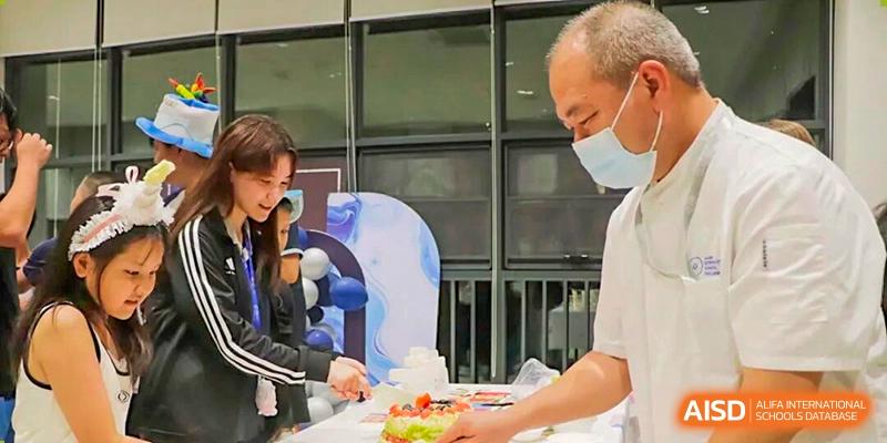 A student is cutting a cake on a table with an older man wearing a mask nearby during a celebration at BASIS International School Park Lane Harbour.