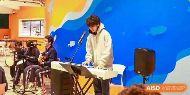 A student in a white hoodie plays a keyboard synthesizer on a brightly lit stage during a performance at BASIS International School Park Lane Harbour.