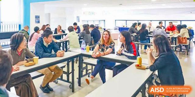 A group of students and a teacher having a casual discussion while sitting at a table in the brightly lit school cafeteria at BASIS International School Park Lane Harbour.