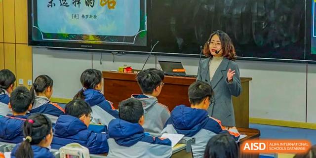 A female teacher standing by a projector screen, smiling and interacting with students seated at desks in the foreground at Nanjing Foreign Language School.