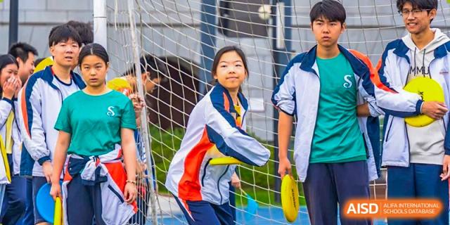 A smiling student is playing ultimate frisbee outdoors, standing near a net with teammates and spectators blurred in the background, showcasing school sports at Nanjing Foreign Language School.