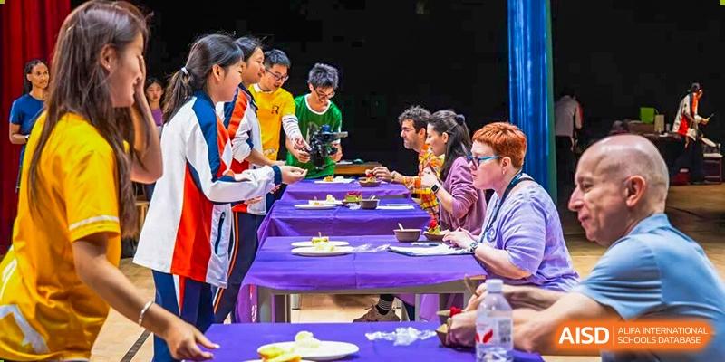 Students and adults are gathered around long tables covered with purple cloths, eating and socializing during a group meal or event, showing community life at Nanjing Foreign Language School.