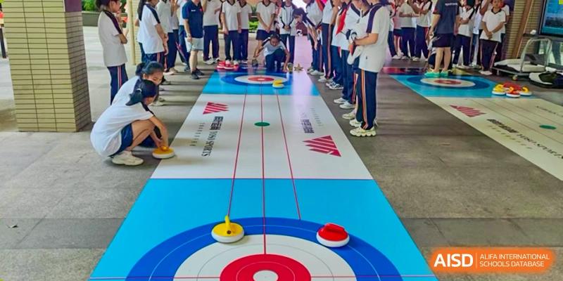 Students are playing a floor curling game on a long mat with blue and red rings, standing in a line in a gymnasium setting, demonstrating school sports at Nanjing Foreign Language School.