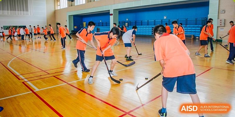 Students in orange and black uniforms are playing a field hockey or floorball game on an indoor court using sticks and a ball, highlighting teamwork and sports at Nanjing Foreign Language School.