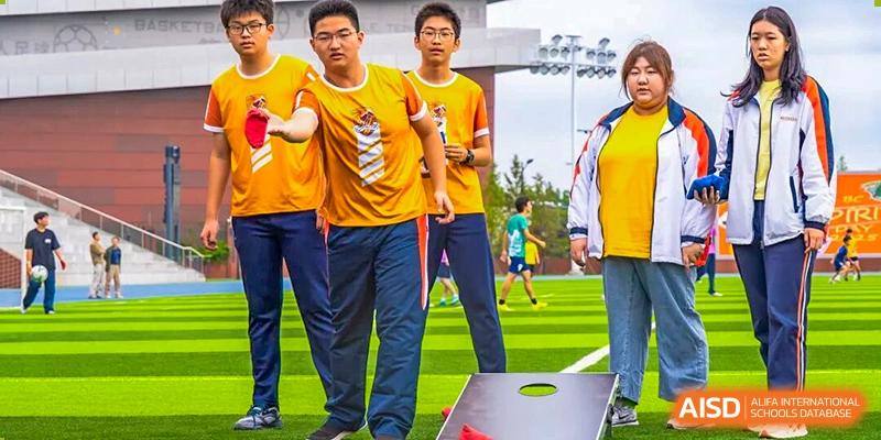 A group of people wearing bright yellow and orange shirts are standing next to a bean bag toss (cornhole) game outdoors during a school sports festival or event at Nanjing Foreign Language School.