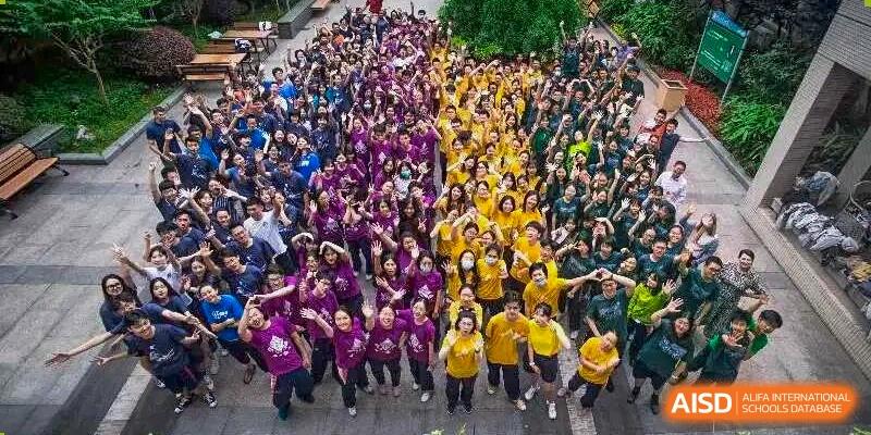 A large crowd of students is gathered outdoors, forming a dense sea of purple and yellow shirts with their arms raised in a celebration or pep rally for a school event at Nanjing Foreign Language School.