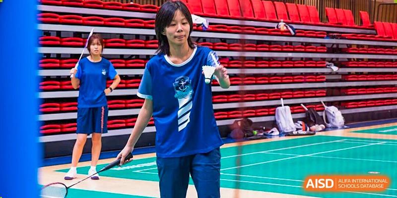 A female student in a blue and white sports uniform is concentrating while holding a racket on a badminton court with red bleachers in the background, promoting school sports at Nanjing Foreign Language School.