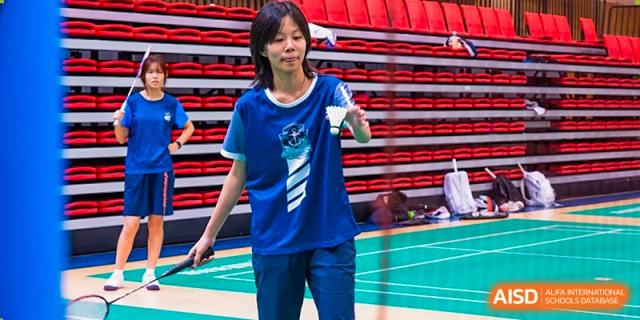 A female student in a blue and white sports uniform is concentrating while holding a racket on a badminton court with red bleachers in the background, promoting school sports at Nanjing Foreign Language School.