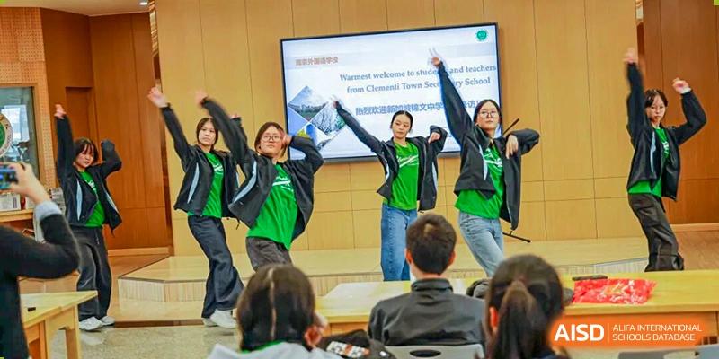A group of students in green and black apparel are performing a lively dance or presentation in front of an audience and a screen in a classroom, illustrating student activities at Nanjing Foreign Language School.