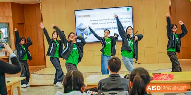 A group of students in green and black apparel are performing a lively dance or presentation in front of an audience and a screen in a classroom, illustrating student activities at Nanjing Foreign Language School.