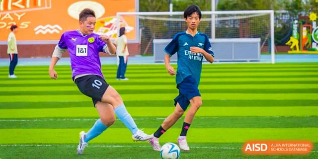 Two young male students playing soccer vigorously on a green field, one in a blue jersey, kicking the ball at Nanjing Foreign Language School.