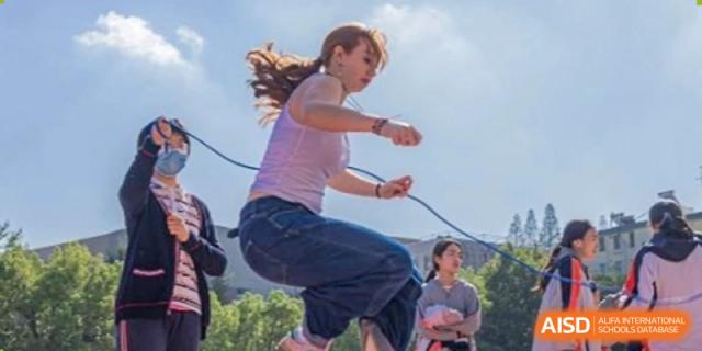 A female student in a white top and wide jeans jumping high in the air outdoors, mid-action during a sports event at Nanjing Foreign Language School.