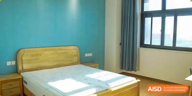 A simple, neat bedroom with light blue walls and a wooden bed frame, possibly a student dorm or staff lodging provided by Nanjing Foreign Language School.
