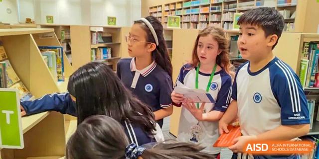Two female students in school uniforms stand in a library and discuss a project while holding printed papers at BASIS International School Shenzhen.