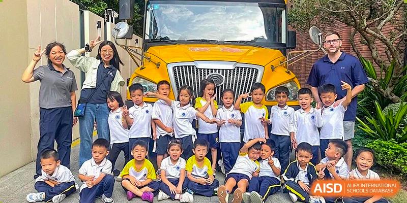 A large group of young primary students in school uniforms pose and smile together in front of a yellow school bus at BASIS International School Shenzhen.