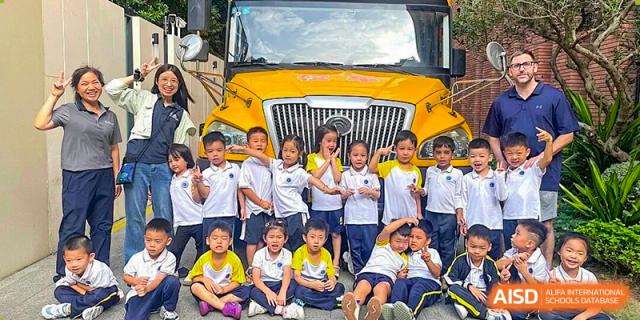 A large group of young primary students in school uniforms pose and smile together in front of a yellow school bus at BASIS International School Shenzhen.