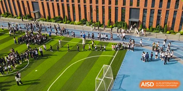 An aerial view of students lined up on a green turf sports field next to a brick building at BASIS International School Shenzhen.