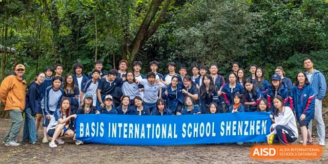 A large group of students poses together behind a blue school banner in a wooded outdoor area at BASIS International School Shenzhen.