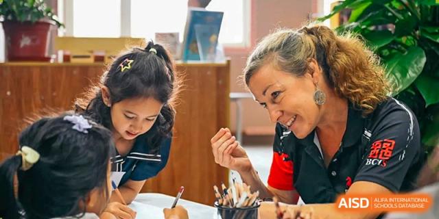 A smiling teacher leans in to provide one-on-one guidance and support to a young student working on a drawing project at Beijing City International School.