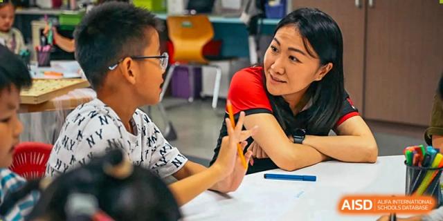 An educator engages in a friendly conversation with a young student while sitting together at a table in a bright classroom at Beijing City International School.