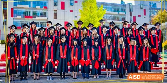 A large group of graduating students in black academic gowns and red sashes poses proudly for a formal commencement portrait outdoors at Beijing City International School.
