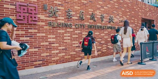 A student with a red backpack walks past a large red brick wall featuring the prominent gold lettering of the main campus entrance at Beijing City International School.