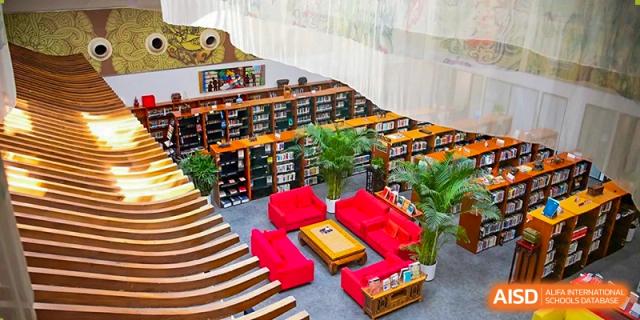 An elevated view shows the modern school library with high wooden bookshelves and a cozy central seating area with red sofas and green plants at Beijing City International School.