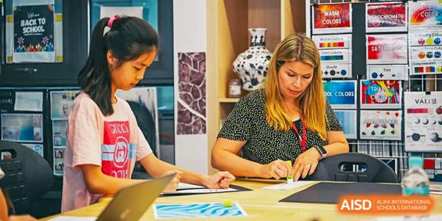 A teacher wearing a floral shirt provides one-on-one guidance to a focused student during a creative art or design lesson in a bright studio at Beijing City International School.