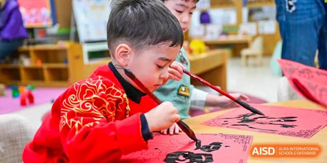 A young student in a traditional red garment carefully practices Chinese calligraphy using a brush and black ink during a cultural lesson at Beijing City International School.