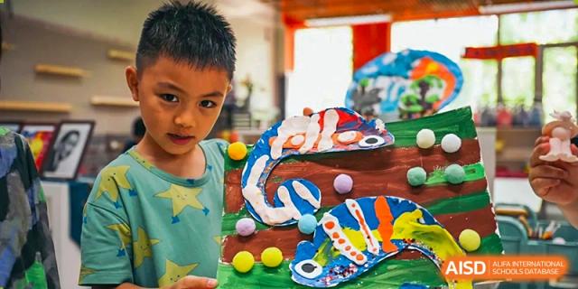A young student proudly displays a vibrant, multi-colored fish artwork decorated with pom-poms that they created during a creative arts and crafts session at Beijing City International School.