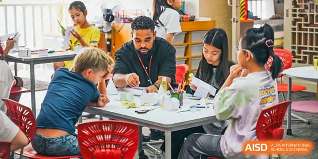 A teacher and a group of students sit around a table and collaborate on a hands-on project involving paper folding and crafting in a modern classroom at Beijing City International School.