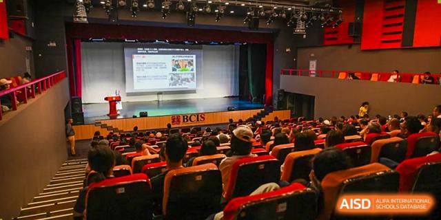 An audience of students and parents fills a large, modern auditorium with red seats to watch a presentation displayed on a professional stage at Beijing City International School.