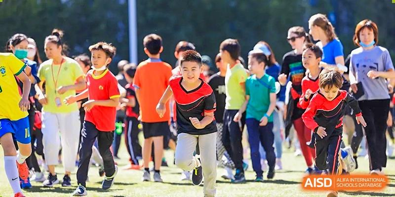 A group of young children in colorful athletic shirts participate in a high-energy outdoor running race on a sunny day at Beijing City International School.