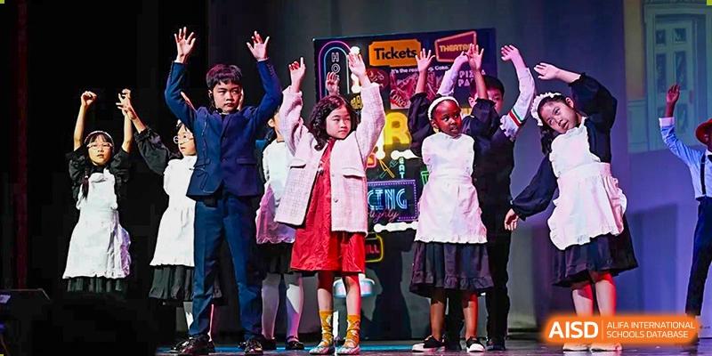 Four young students in formal costumes perform a choreographed theatrical play on a professional stage with colorful backdrop lights at Beijing City International School.