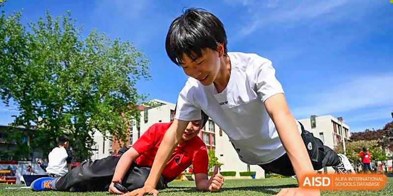 Two male students in red and white shirts perform focused push-ups on a green athletic field during an outdoor physical fitness session at Beijing City International School.
