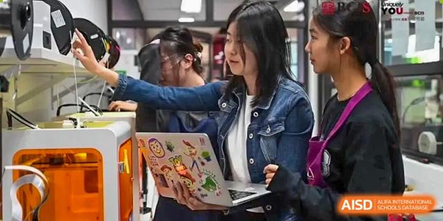 Two female students collaborate on a project while looking at a laptop decorated with various colorful stickers in a modern classroom at Beijing City International School.