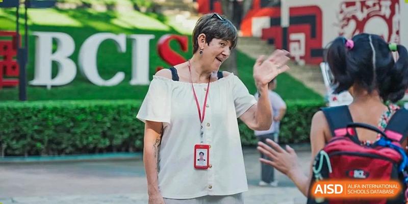 A smiling staff member wearing a school identification lanyard waves to someone while standing in front of a large green CIS sign at Beijing City International School.