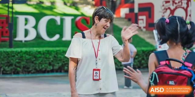 A smiling staff member wearing a school identification lanyard waves to someone while standing in front of a large green CIS sign at Beijing City International School.