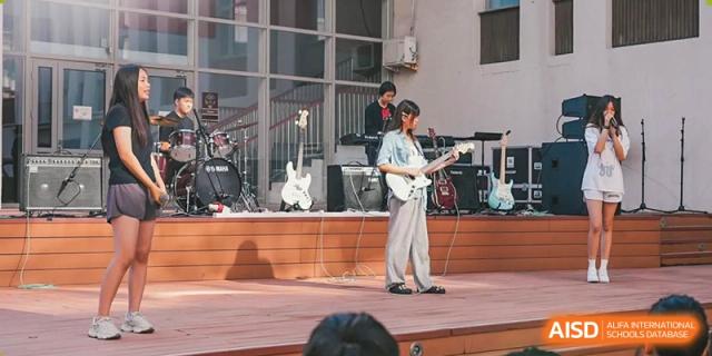 A student guitarist performs a live solo on an outdoor wooden stage during a school music festival while a drummer plays in the background at Beijing City International School.