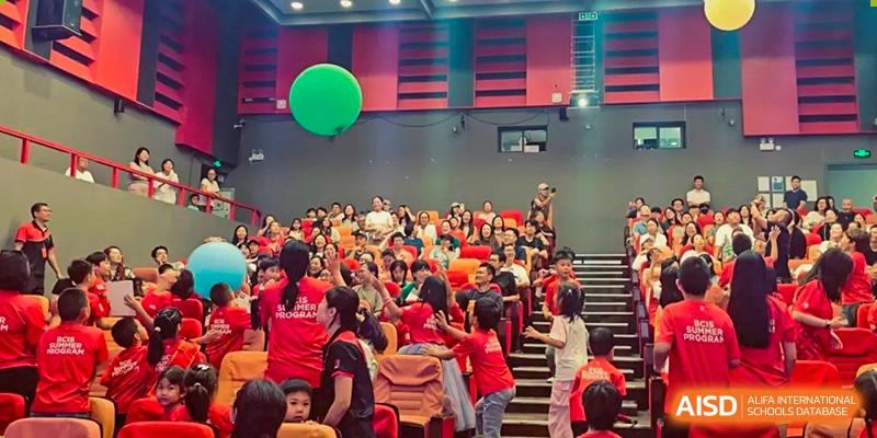 Students in red shirts gather in a large auditorium and reach up toward large green and blue balloons floating above the audience during a lively event at Beijing City International School.