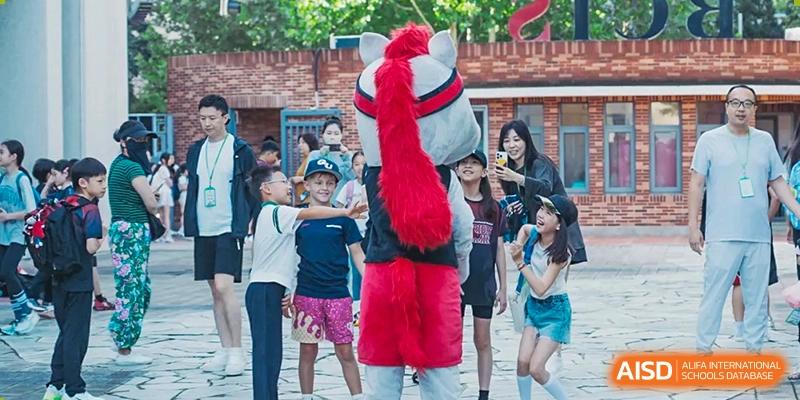 Young students interact excitedly with a person wearing a large grey and red horse mascot costume in an outdoor courtyard during a festive campus celebration at Beijing City International School.
